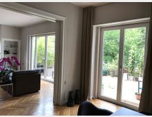 Wohnraum mit hellem Parkettboden, großen bodentiefen Fenstern, die einen Blick auf eine bepflanzte Terrasse bieten. Im Vordergrund stehen eine dunkle Couch, ein Tisch mit einer Orchidee und Bücherregale im Hintergrund.