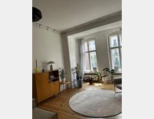 Wohnzimmer mit Holzfußboden, einem runden, hellen Teppich und großen Fenstern mit weißen Vorhängen. Links steht eine Holzkommode mit Dekoration, und mehrere Zimmerpflanzen sind im Raum verteilt. Stuckverzierungen sind an der Decke sichtbar.