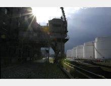 Industriegebiet mit alten Backsteingebäuden, einem Kran und Gleisen auf Kopfsteinpflaster im Vordergrund; im Hintergrund stehen mehrere große, weiße Tanks neben einem Wasserkanal.