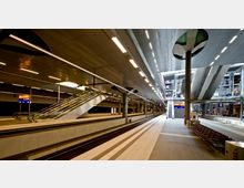 Moderne Bahnhofshalle mit beleuchteter Decke, zwei Bahnsteigen und Gleisen. Rolltreppen und Treppen führen zu den oberen Ebenen, während Sitzbänke und Beschilderungen entlang der Bahnsteige sichtbar sind.