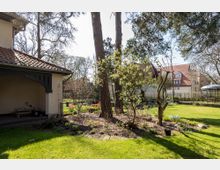 Wohnhausgarten mit gepflegter Rasenfläche, Blumenbeeten und Bäumen im Vordergrund. Links ein Gebäude mit roten Dachziegeln und überdachter Terrasse, im Hintergrund weitere Wohnhäuser und ein Pavillon.
