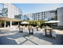 Moderner Außenbereich eines Gebäudekomplexes mit Glasfassaden und Rundformen. Im Vordergrund stehen Holztische mit weißen Hockern sowie Sonnenschirme auf einem gepflasterten Platz, im Hintergrund eine dichte Hecke.