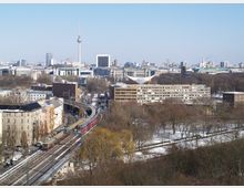 Stadtansicht von Berlin mit Blick auf den Fernsehturm in der Ferne, das Reichstagsgebäude mit gläserner Kuppel und umliegenden modernen und historischen Gebäuden. Im Vordergrund verlaufen schneebedeckte Bahngleise mit vorbeifahrenden Zügen sowie ein Park mit kahlen Bäumen.