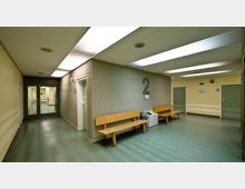 Ein schlicht gehaltener Innenbereich eines Gebäudes, vermutlich ein Wartebereich in einer Klinik oder einem öffentlichen Gebäude. Die Wände sind hellgelb gestrichen, der Boden besteht aus grünem Linoleum, es gibt Holzbänke, Mülleimer und Türen; die Wand ist mit einer großen "2" markiert.