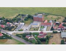 Luftaufnahme eines Dorfes mit einer Hauptstraße, umgeben von Wohnhäusern, Höfen und landwirtschaftlichen Flächen. In der Mitte des Bildes steht ein großes, mehrstöckiges Ziegelgebäude sowie ein gelbes, einstöckiges Gebäude, beide auf einem umzäunten Grundstück.