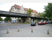 Straßenszene in einer städtischen Umgebung mit gepflastertem Gehweg im Vordergrund, einer Hauptstraße mit parkenden und fahrenden Autos sowie einer erhöhten Eisenbahnbrücke aus Stahl. Im Hintergrund befinden sich mehrstöckige Altbauten mit roten Ziegeldächern und detaillierten Fassaden.