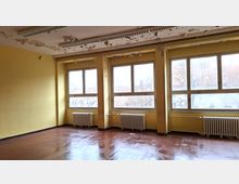 Leerer, renovierungsbedürftiger Innenraum mit gelb gestrichenen Wänden, großen Fensterreihen und Heizkörpern darunter. An der Decke hängen Leuchtstofflampen, und die Farbe blättert großflächig ab; der Boden ist glatt und dunkel.
