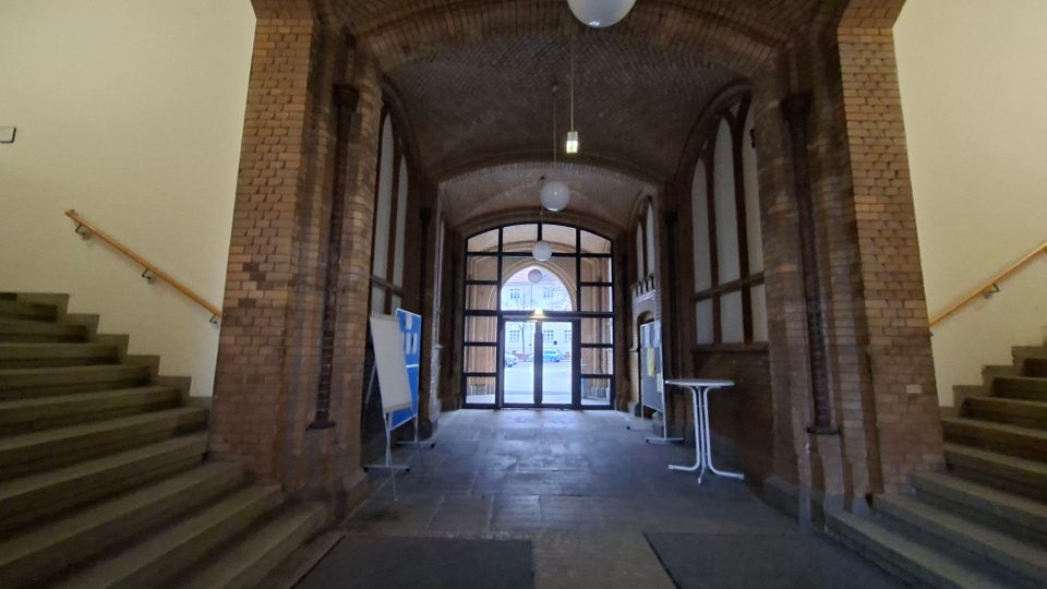 Gewölbter Durchgang in einem Backsteingebäude mit Tonnengewölbe, Rundbogenöffnungen und hängenden Kugelleuchten. Links und rechts führen Treppen mit Holzhandläufen nach oben; am Ende steht eine verglaste Türanlage mit Blick nach draußen.