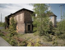 Ehemaliges Bahnhofsgebäude aus Backstein mit teilweise zerbrochenen Fenstern, umgeben von wildem Bewuchs und kleinen Bäumen, neben alten Bahngleisen. Im Hintergrund ist ein weiteres, schlichtes Gebäude sowie ein Strommast zu sehen.