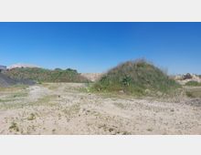 Eine offene Kies- und Sandfläche mit einzelnen, mit Gras und Sträuchern bewachsenen Erdhügeln im Hintergrund. Der Himmel ist klar und blau, und links ist ein Gebäudeausschnitt erkennbar.