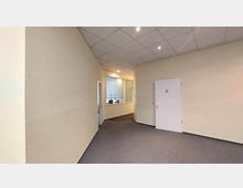 Ein schlichter Büroflur mit hellgelben Wänden, grauem Teppichboden und einer abgehängten Decke mit eingebauten Spotlights. Rechts ist eine weiße Tür mit Rollstuhlsymbol, links ein Bereich mit Fenstern, die in einen Besprechungsraum blicken.