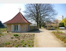 Kleines Backsteingebäude mit rotem Ziegeldach und Fensterläden, umgeben von einem Gartenbereich mit kahlen Beeten und Wegen. Im Hintergrund steht ein großer, unbelaubter Baum, und ein modernes Hochhaus ist am Horizont erkennbar.
