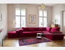 Wohnzimmer mit hellen Wänden, großen Sprossenfenstern und einer roten Ecksofa-Garnitur. Ein bunter Teppich bedeckt den Holzparkettboden, während ein kleiner runder Beistelltisch sowie eine Kommode mit Lampe und dekorativen Elementen den Raum akzentuieren.