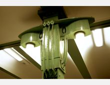 Grünes, kunstvoll verziertes Stahlträger-Element einer Bahnhofs- oder U-Bahn-Architektur, mit integrierten Leuchten und sichtbaren Deckenkonstruktionen sowie modernen Deckenlampen im Hintergrund.