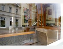 Reflexion in der Glasfront eines Cafés, das den Blick auf eine gepflasterte Straße mit angrenzenden Gebäuden und Geschäften zeigt. Im Inneren des Cafés sind ein Korbstuhl, ein Tisch und an der Wand hängende Bilder zu erkennen.