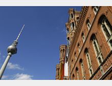 Der Fernsehturm in Berlin, links im Bild, ragt in den blauen Himmel, während rechts die rote Backsteinfassade des historischen Roten Rathauses mit dekorativen Fensterelementen und drei wehenden Fahnen zu sehen ist.