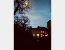 Historisches Gebäude mit rotem Ziegeldach und erleuchteten Fenstern in einer nächtlichen Szenerie, umgeben von Bäumen und einem wolkigen Himmel, durch den der Mond scheint.