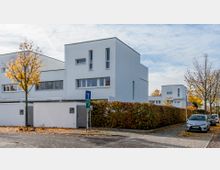 Moderne Wohnsiedlung mit weißen, kubischen Gebäuden und flachen Dächern, umgeben von Hecken und herbstlich gefärbten Bäumen. Im Vordergrund ist eine Straßenecke mit einem Straßenschild sowie parkenden Autos zu sehen.