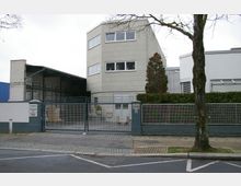 Industrielles Gebäude mit einer hellgrauen Fassade und mehreren Fenstern, umgeben von einer niedrigen Mauer mit einem Metalltor. Im Hof sind eine überdachte Lagerfläche sowie ein Baum und Heckenpflanzen sichtbar.