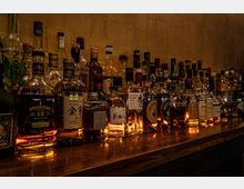 Regal einer Bar mit einer großen Auswahl an Spirituosenflaschen, darunter Whisky, Rum und andere alkoholische Getränke, auf einer hölzernen Theke vor einer schlichten, beigefarbenen Wand. Die Flaschen sind eng aneinandergereiht, teilweise mit leuchtenden Etiketten und reflektierenden Glasoberflächen.