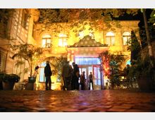 Historisches Gebäude mit heller Fassade und Rundbogenfenstern, beleuchtet durch bunte Lichteffekte, umgeben von Palmen und Pflanzen. Vor dem Eingangsbereich stehen Menschen auf einem gepflasterten Hof.