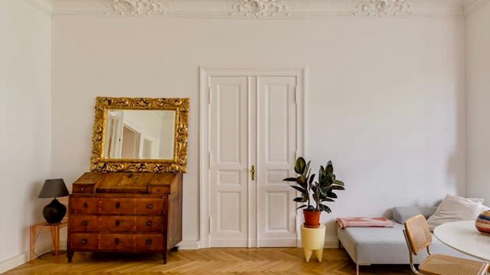 Heller Wohnraum mit weißen Wänden, Stuckdecke und Doppelflügeltür in der Mitte. Links stehen eine antike Holzkommode mit großem goldgerahmtem Spiegel und eine kleine Lampe, rechts ein graues Sofa neben einer Topfpflanze auf Fischgrätparkett.