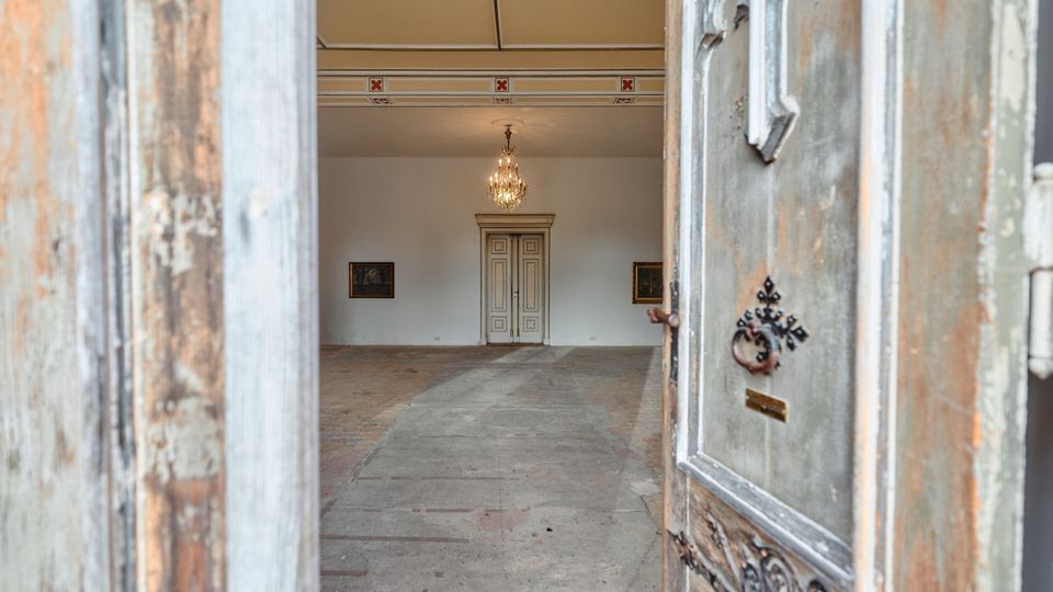 Blick durch eine halb geöffnete, verwitterte Holztür in einen hohen, hellen Innenraum mit Stuckdecke und Kronleuchter. Im Hintergrund steht eine doppelflügelige Tür an der Stirnwand, flankiert von zwei gerahmten Bildern.