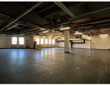 Industriehalle mit Betonboden, sichtbaren Lüftungsrohren an der Decke und weißen Wänden, teils unterteilt durch Säulen. Links sind mehrere Fenster mit Gitterstäben und eine Metalltür zu sehen, während der Raum insgesamt spärlich beleuchtet ist.