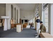 Eleganter Innenraum eines Modegeschäfts mit modernen, geschwungenen Elementen. Zu sehen sind Schaufensterpuppen in Kleidung, gebogene beige Möbel, dekorative Bögen, große Fenster mit Tageslicht und minimalistische Präsentationsflächen.