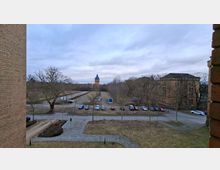 Blick von einem Backsteingebäude auf ein Gelände mit gepflasterten Wegen, einer Wiese und einem Parkplatz mit mehreren Autos. Im Hintergrund stehen laublose Bäume, ein hoher Turm mit spitzem Dach und ein großes historisches Backsteingebäude unter bewölktem Himmel.