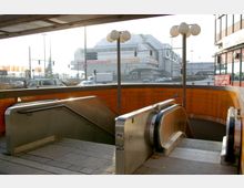 U-Bahn-Zugang mit Rolltreppe und Treppe, umgeben von orangefarbenen Fliesenwänden, im Vordergrund. Im Hintergrund ist eine stark befahrene Straße und ein großes, modernes Gebäude mit Glas- und Metallfassade zu sehen.