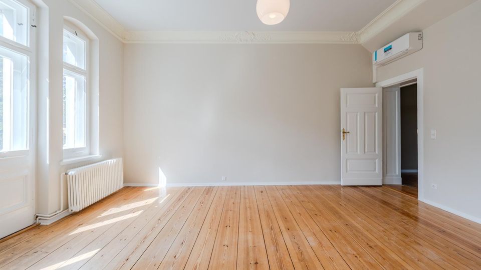 Heller, leerer Altbau-Raum mit breiten Holzdielen, hohen Decken mit Stuck und einer runden Deckenleuchte. Links große Sprossenfenster über einem Heizkörper, rechts eine offene Tür zum Nebenraum und eine wandmontierte Klimaanlage.