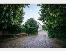 Freistehendes weißes Gebäude mit rotem Giebeldach, umgeben von dichter Vegetation. Der Vordergrund zeigt einen gepflasterten Weg, flankiert von Bäumen und einer mit Efeu bewachsenen Mauer.