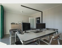 Moderner Büroraum mit grauen Wänden, einem großen Schreibtisch aus Metall, zwei Monitoren, einer Tastatur und einem Telefon. Im Hintergrund stehen ein metallisches Sideboard mit Deko und ein schwarzes Ledersofa, daneben eine Glastür und eine Stehlampe.