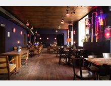 Ein gemütliches Café oder Restaurant mit Holzboden, violett gestrichenen Wänden und gedämpfter Beleuchtung. Die Einrichtung besteht aus Holztischen und -stühlen sowie gepolsterten Sesseln, während dekorative Lampen und ein Rohrsystem an der Wand die Atmosphäre prägen.