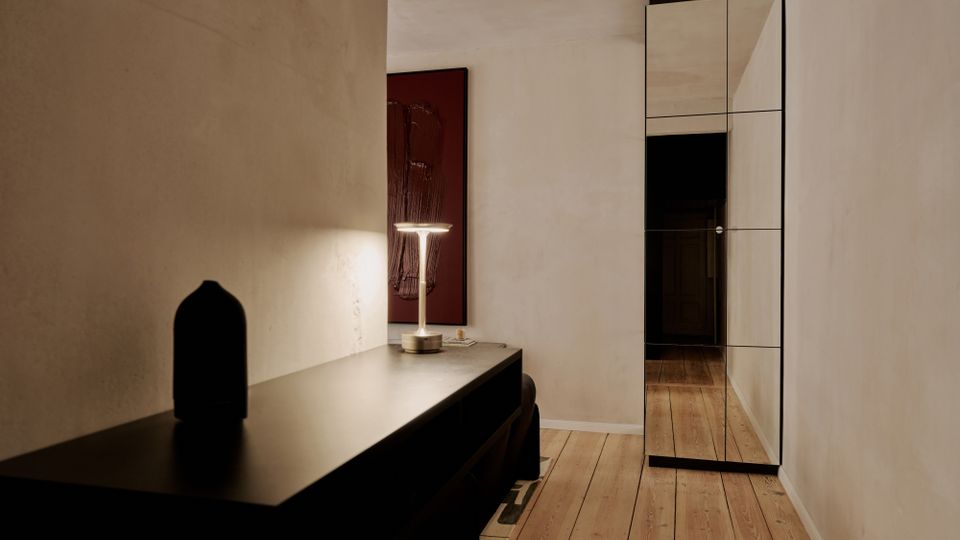 Schmaler, moderner Flur mit hellem Putz an den Wänden und breiten Holzdielen am Boden. Links steht eine dunkle Konsole mit Tischlampe und Wandbild, rechts ein hoher Spiegel mit schwarzem Rahmen vor dem Durchgang in einen dunkleren Raum.