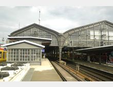 Großer Bahnhof mit Glasdach und Stahlkonstruktion, mehrere Bahngleise und Bahnsteige, ein kleineres Gebäude mit verglasten Fenstern im Vordergrund; im Hintergrund ist ein Fernsehturm sichtbar.