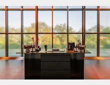 Ein eleganter Tisch in einem modernen, lichtdurchfluteten Raum mit großen, bodentiefen Fenstern. Auf dem Tisch stehen Weinflaschen, Metallkühler und dekorative Elemente, im Hintergrund sind Bäume durch die Fenster sichtbar.