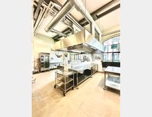 Industrieküche mit Edelstahl-Arbeitsflächen, einem großen Lüftungssystem an der Decke und Fliesenboden. Im Hintergrund sind Lagerregale, ein Kühlschrank und große Fenster, die natürliches Licht hereinlassen.