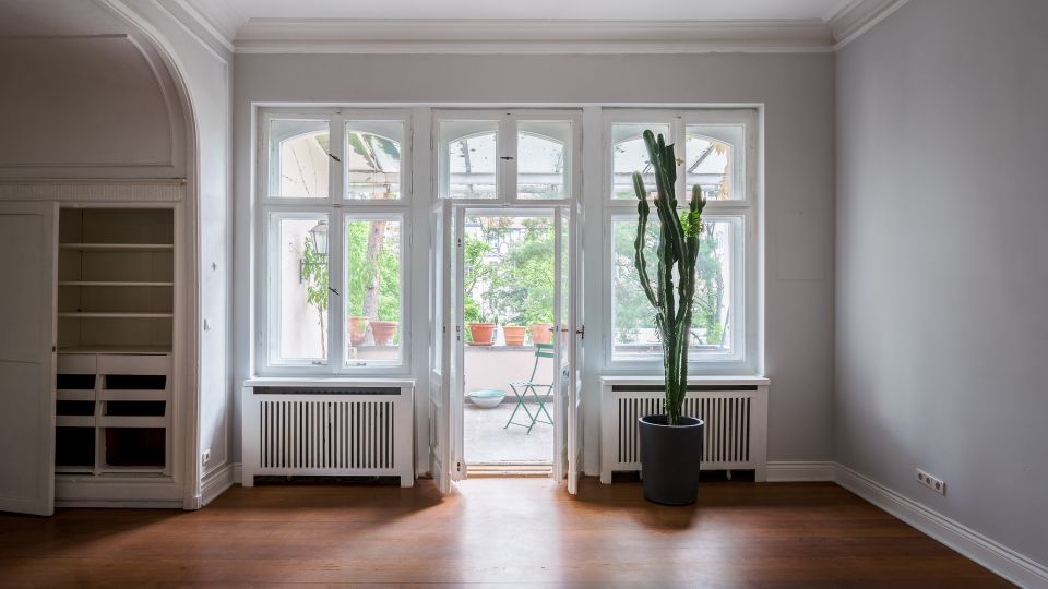 Heller, leerer Wohnraum mit hohen Decken, Stuckleisten und großem dreiteiligem Fenster mit Balkontür zur Terrasse. Links ein Einbauschrank in einer Nische, unter den Fenstern Heizkörperverkleidungen und rechts ein großer Kaktus im Topf auf Holzfußboden.