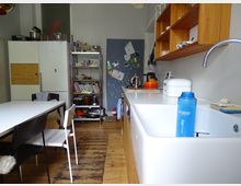 Kleine Küche mit Holzboden und einer weißen Arbeitsplatte, darauf eine Spüle und verschiedene Küchenutensilien. An der Wand befinden sich offene Holzregale, ein Regal aus Metall mit Büchern und Geschirr sowie ein großer Kühlschrank; im Hintergrund eine Pinnwand mit Notizen und Zeichnungen.