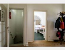 Ein beleuchteter Flur in einem Wohnraum mit Holzboden; links führt eine Tür zu einer Treppe, rechts ist eine Garderobe mit Jacken und Taschen sichtbar. Geradeaus öffnet sich ein Durchgang zu einem Essbereich mit Sitzbank, Tisch und heller gewölbter Decke.