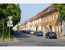 Straßenszene mit Ziegelhäusern und roten Dächern auf der rechten Seite, daneben parkende Autos entlang der Straße. Links ein Bürgersteig mit Straßenlaternen und Bäumen, Verkehrszeichen und ein Tempo-30-Schild sichtbar.