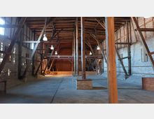 Großer Innenraum einer historischen Scheune oder Lagerhalle mit sichtbaren Holzbalken, freiliegenden Backsteinmauern und einem Betonboden. Die Decke ist hoch, und Hängeleuchten beleuchten den Raum, der vorne eine Bühne mit hölzernem Geländer besitzt.