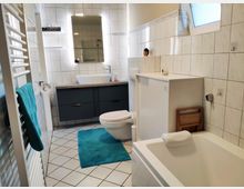 Modernes Badezimmer mit weißen Fliesenwänden und gefliesten Boden in hellen Farben. Zu sehen sind eine Badewanne, ein Wand-WC, ein dunkelgrauer Waschtisch mit Waschbecken und beleuchtetem Spiegel darüber sowie ein Heizkörperhandtuchhalter mit einem blauen Handtuch.