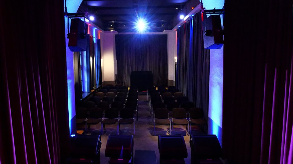 Blick von der Bühne in einen kleinen Veranstaltungssaal mit Reihen aus dunklen Stühlen und einem Mittelgang. Dunkle Vorhänge, seitliche Säulen und blau-violette Bühnenbeleuchtung sowie Lautsprecheranlagen prägen den Raum.