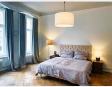 Ein Schlafzimmer mit einem weiß gepolsterten Bettkopfteil, einem Doppelbett mit heller Bettwäsche und zwei Nachttischen mit Lampen. Der Raum verfügt über zwei große Fenster mit grauen Vorhängen, eine Holzdielenboden und eine Wanduhr links.