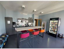 Kleine Teeküche mit grauen Wänden und dunklem Boden, ausgestattet mit einer Küchenzeile mit Kaffeemaschinen, Mikrowellen und Schränken. In der Mitte steht ein hoher Tisch mit bunten Hockern, rechts ein Getränke- und Snackautomat, links mehrere Mülltrennungsbehälter.