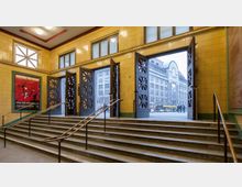 Treppenhaus eines historischen Gebäudes mit gelb gekachelten Wänden, Metallhandläufen und großen Holztüren, die nach außen auf einen Platz mit einem gegenüberliegenden Gebäude im klassischen Stil führen. Ein rotes Werbeplakat und Fenster mit Milchglas zieren die Wände.