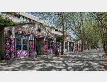 Straßenszene mit kleinen asiatischen Restaurants und Geschäften, erkennbar an bunten Schildern und Menüs vor den Eingängen. Der Bürgersteig ist von Bäumen gesäumt, deren Schatten auf den Boden fällt, und im Hintergrund sind Passanten unterwegs.